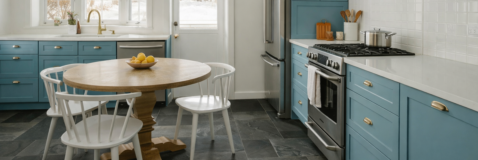 Modern kitchen featuring blue cabinets, stainless steel appliances, and a wooden round table with a bowl of lemons, illustrating cabinet painting transformation by Queen City Painting & Decorating.