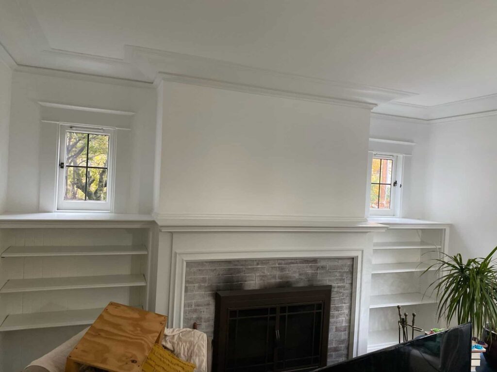 Interior room featuring freshly painted white walls, a fireplace with a brick surround, built-in shelves, and natural light from windows, illustrating the transformative effect of interior house painting.